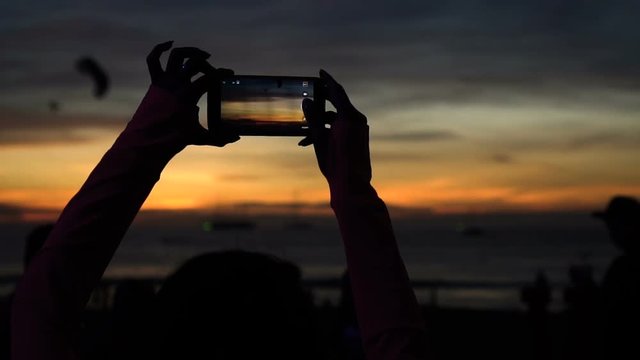 Silhouette of woman taking picture of sunset with phone at beach - video in slow motion