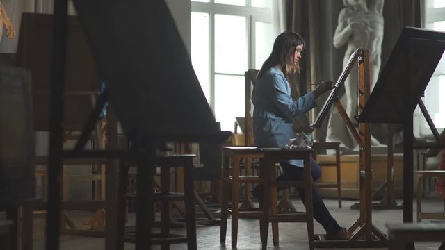 Young woman painter in blue dress draws behind an easel in a light and spacious art studio.General plan: the artist's workshop with interesting creative objects of the interior.