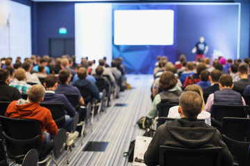 Business and entrepreneurship symposium. Speaker giving a talk at business meeting. Audience in the conference hall. Rear view of unrecognized participant in audience.