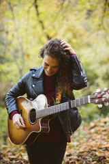 Fototapeta na wymiar Portrait of beautiful smiling woman playing guitar on forest, fashion lifestyle. Girl wearing black jacket.