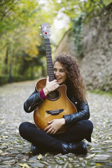 Beautiful young woman with a guitar sitting on the forest. Girl wearing black jacket, fashion lifestyle.