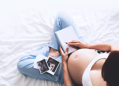 Pregnant woman makes notes in notebook and holding ultrasound image