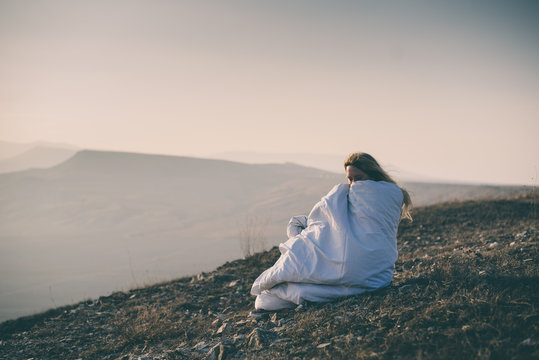 Lonely sad and crying young woman sitting wrapped in blanket on cliff of mountain. Toned image