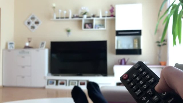 Person sitzt im Wohnzimmer auf Sofa und schaltet Fernseher an bzw. aus