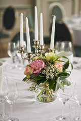 Little bouquet of pink peonies and orange roses stands before candleholder on the dinner table