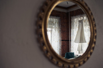 Eleganr wedding dress hangs on the tall window and reflects in the mirror