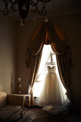 White wedding dress hangs before the window in luxury room