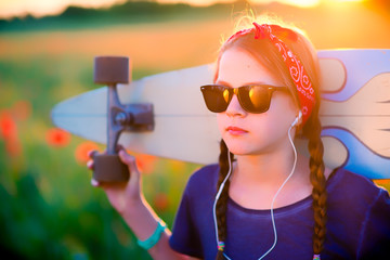 Young hipster girl with braids in sunglasses and a red sash on her head, listening to music with a longboard on her shoulders - against a summer meadow at sunset