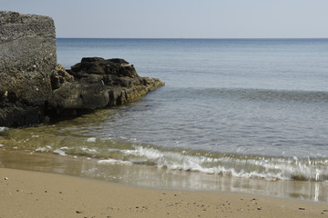 Waves comes in to a sandy beach and a cliff to the left with horizon and a blue sky in background