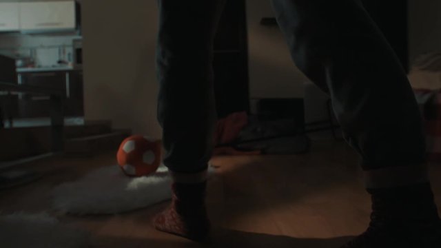 Legs of unrecognizable person in red patterned socks and grey sweat pants walks across living room with pile of clothes on floor, kicks football, close up slow motion tracking shot