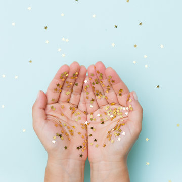 Top View On Woman's Hands Covered With Golden Stars Confetti On Light Blue Background. Christmas And Celebration Concept