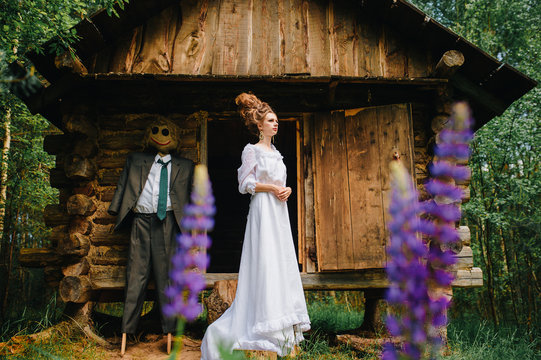 Young beautiful skinny bride girl in vintage wedding dress and hairstyle standing beyond old wooden house in forest with scarecrow from fairytale. Classical russian fable concept and idea. Sorcery.