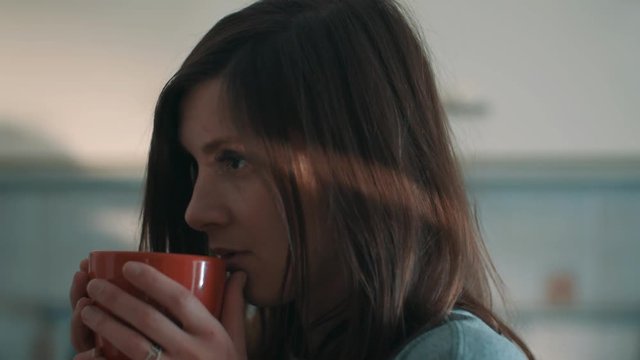 Young attractive brunette caucasian girl drinking coffee out red mug and winces in kitchen at early morning, beam of light across face, close up slow motion