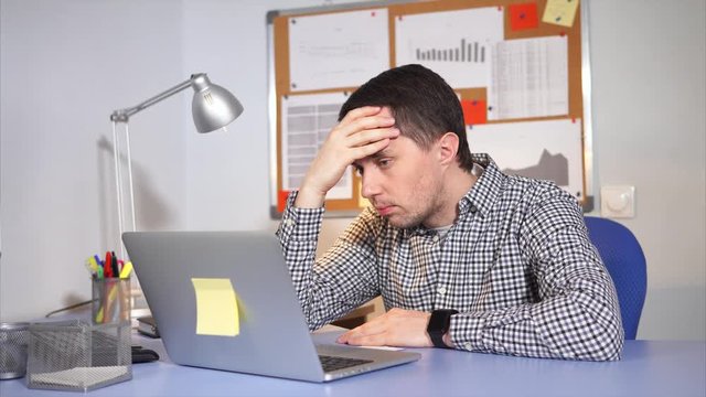 A man who looks tired keeps his head, he prints the text on the laptop keyboard to complete the office project and send via the Internet to the chief's mail