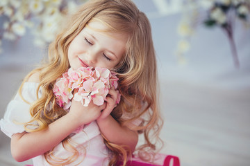 Portrait of a smiling little girl in beautiful dress sitting on a light wooden floor .Cute girl holding a flower in his hand and presses it to his face with my eyes closed