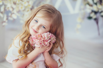 Smiling little girl in beautiful dress sitting on a light wooden floor .Cute girl holding a flower in his hand and presses it to his face.