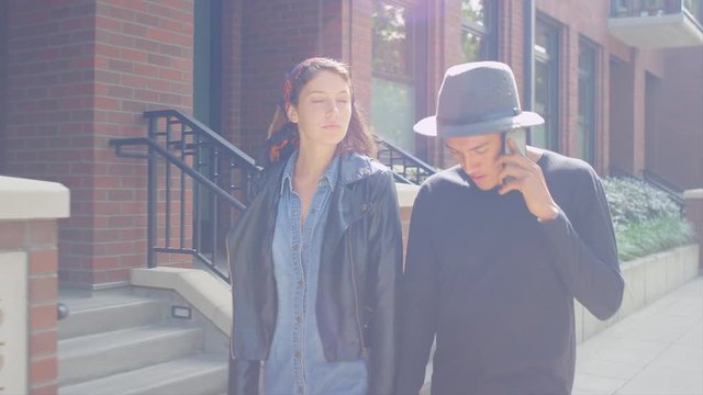 A hipster couple walking down the sidewalk arm in arm, boyfriend talking on his phone