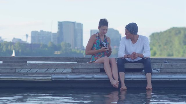 A couple sits on dock at a lake and takes photos with an old camera, city in the background