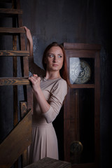 Beautiful girl in dark room with wooden staircase and vintage clock. Wore in beautiful dress, good looks, model elegant red haired, studio shot