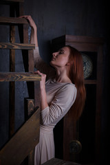 Beautiful girl in dark room with wooden staircase and vintage clock. Wore in beautiful dress, good looks, model elegant red haired, studio shot