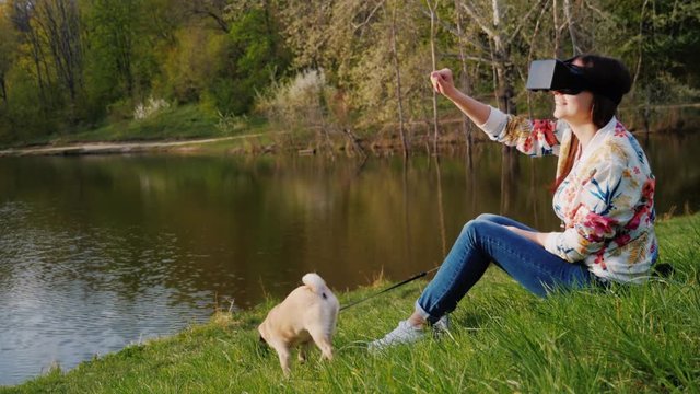 Young woman in the virtual reality. Sits in a picturesque place on the lake, next to her dog pug breed