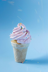 Cupcake in an ice-cream cone - perfect summer dessert for an outdoor birthday party. Strawberry buttercream icing contrasting with sky blue background. Blurry lines from colorful sprinkles. 