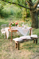 Fototapeta na wymiar picnic, food, summer, holiday concept - festively decorated wooden picnic table among trees, bouquet, pitcher of lemonade, glasses, fruit plates, candlesticks, pillows and blanket on chairs and bench