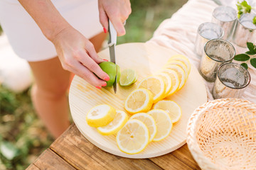 Obraz na płótnie Canvas picnic, people, food, summer, nature concept - young slender girl in a white dress with a knife cut lemon into round slices, lie on a plate of lemons and green lime, dishes, glasses on wooden table