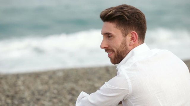 Giovane ragazzo in camicia bianca sorride seduto nella spiaggia in riva al mare. Concetto Estate. 