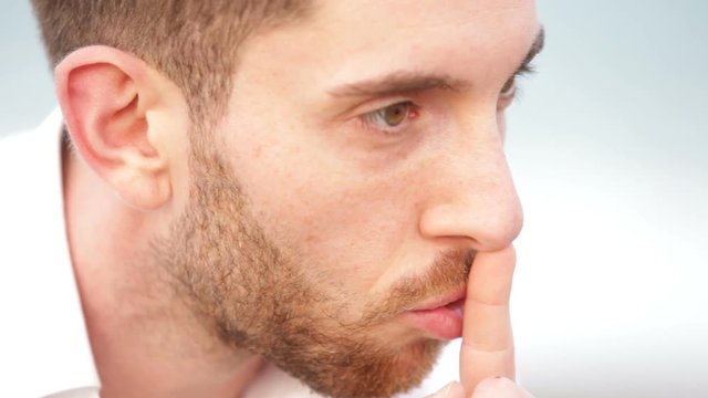 Ragazzo con camicia bianca zittisce con il dito indice sul naso. 