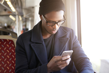 Cute dark-skinned student in stylish spectacles going to University by bus, holding his web-enabled...