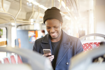 Laughing dark-skinned male in tram. African American using contemporary smartphone sitting in...