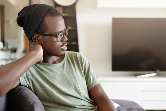 Time to relax. Profile of fashionable dark-skinned student in eyewear and hat sitting at home on comfortable chair or couch, having relaxed carefree look after morning lectures at university