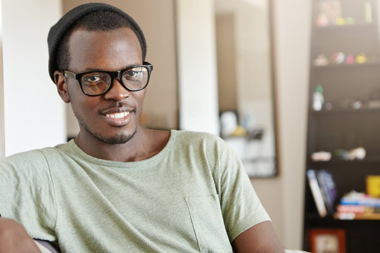 Indoor shot of young Afro American freelancer in hat and rectangular glasses feeling happy and relaxed after he finished work, sitting in his room with cozy interior. Black man resting at home