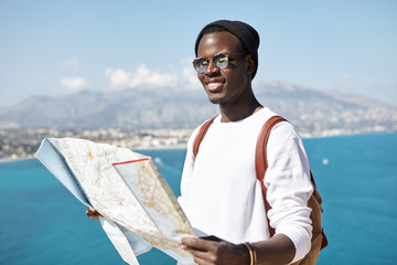 Happy handsome young dark-skinned male traveler standing on top of mountain with paper map above vast ocean and resort town, having joyful look while traveling around the world in company of friends