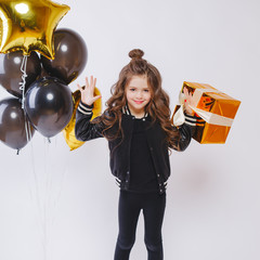 Little modern hipster girl in fashion clothes stand near balloons and hold gold present. Birthday.