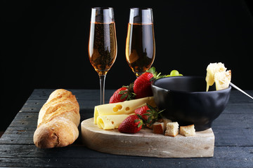 Gourmet Swiss fondue dinner with assorted cheeses on a board and strawberries alongside a heated pot of cheese fondue with two forks dipping bread and white wine behind in a tavern or restaurant