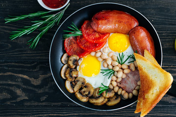 Traditional English breakfast eggs, tomatoes, bacon, sausages, mushrooms, toast and beans in a pan decorated with rosemary and orange juice on a wooden background Top view