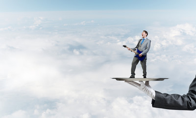 Businessman on metal tray playing electric guitar against blue sky background
