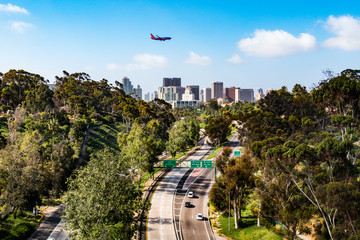 An airplane flies over the Cabrillo Freeway (State Route 163) as it passes through Balboa Park and into the downtown area of San Diego, California.   - obrazy, fototapety, plakaty