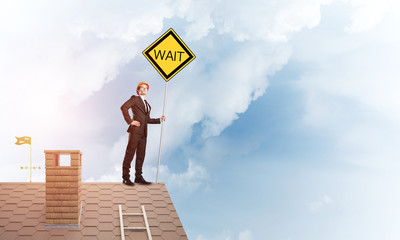 Young businessman on house brick roof holding yellow signboard. Mixed media