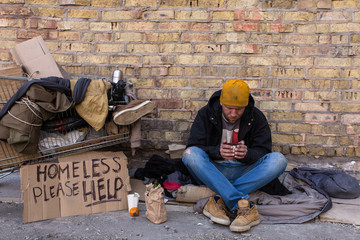Homeless man sitting on the street, near the wall and cart with his stuff. Holding cup of coffee and looking in it.