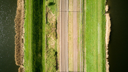 Fototapeta premium Train lines. Vertical aerial drone view of a pair of train tracks bordered by water and pathways creating abstract lined textures.