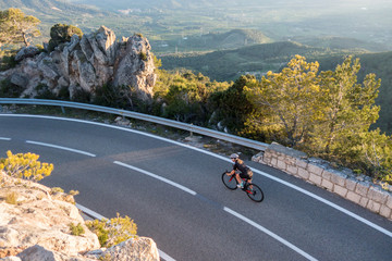 Naklejka premium Young female cyclist riding, cycling on sunset ray glissened road high in the spainish mountians narrow road surrounded by green trees and huge sand colored rocks.
