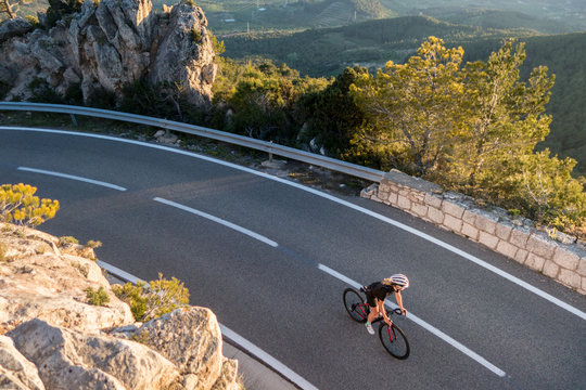 Young female cyclist riding, cycling on sunset ray glissened road high in the spainish mountians narrow road surrounded by green trees and huge sand colored rocks.