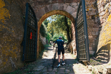 Fototapeta na wymiar Strong male sprinter professional cyclist in midday sunlight, light from above, approaches open gate with iron and tan bricks in a high gothic village in the spanish mountains.
