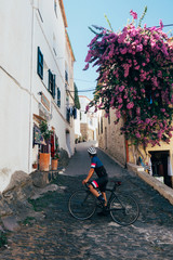 Male strong pro cyclist staring down alley in the village of altea, spain with all buildings painted white with narrow pave brick roads, in colorful kits clothing on a black bicycle with purple tree.
