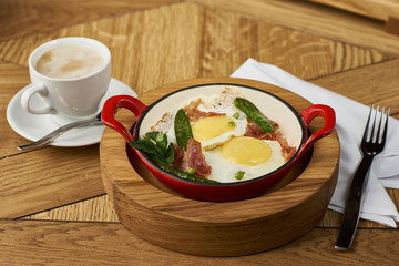 bacon and eggs with green peas in red pan served on wooden table in restaurant