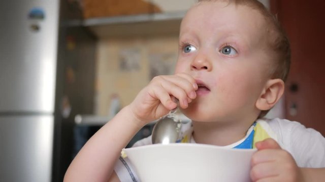 A cute blonde boy eats a porridge with a spoon from a plate of the house. Close-up