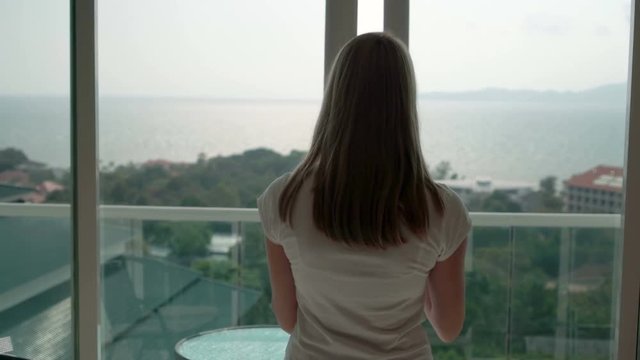 Woman in white t-shirt opening balcony doors and looking out. Enjoying the sea view outside